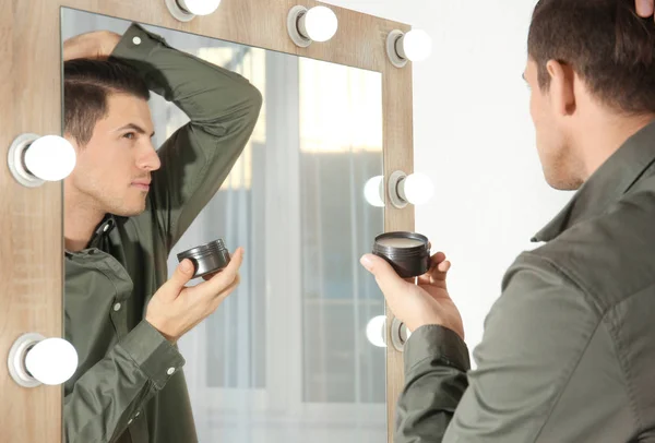 Hombre joven guapo aplicando arcilla para el pelo cerca del espejo en casa —  Fotos de Stock