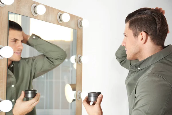 Hombre joven guapo aplicando arcilla para el pelo cerca del espejo en casa —  Fotos de Stock