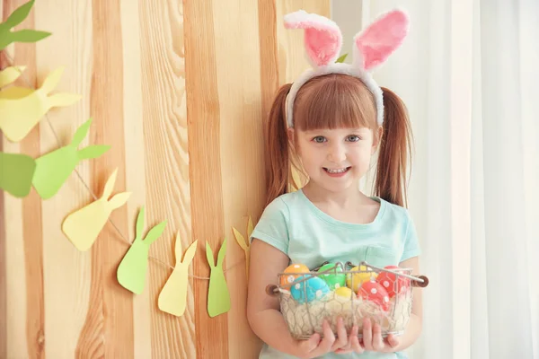 Menina bonito com orelhas de coelho segurando cesta cheia de ovos de Páscoa coloridos dentro de casa — Fotografia de Stock