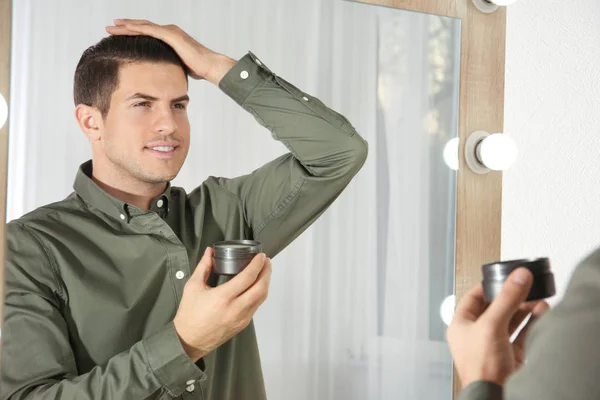 Hombre joven guapo aplicando arcilla para el pelo cerca del espejo en casa —  Fotos de Stock