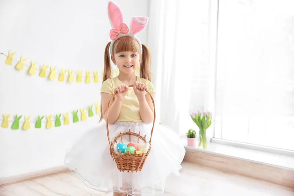 Schattig klein meisje met bunny oren mand vol kleurrijke paaseieren binnenshuis te houden — Stockfoto