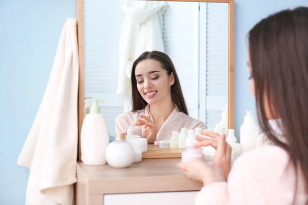 Mujer joven con frasco de crema de manos delante del espejo en el interior — Foto de Stock