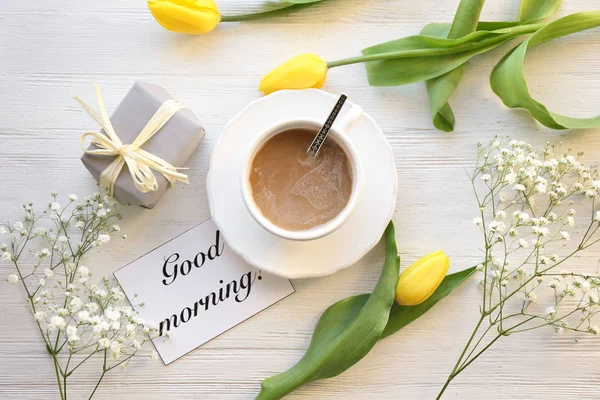 Чашка кофе на завтрак, подарок, цветы и открытка со словами "Доброе утро" на столе, вид сверху — стоковое фото
