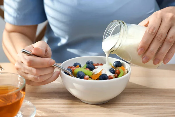 Женщина, наливающая вкусный йогурт на фрукты в миске за столом — стоковое фото