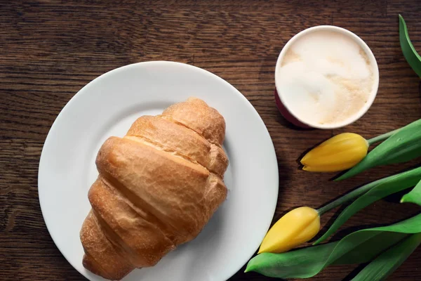 Deska s croissantem, šálek kávy a květiny na stole, pohled shora. Chutná snídaně — Stock fotografie