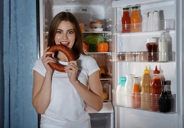 Young woman with sausage standing near refrigerator at night