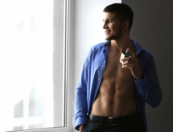 Knappe man in unbuttoned shirt en met fles parfum in de buurt van venster — Stockfoto