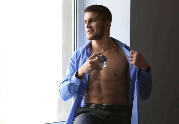 Hombre guapo en camisa desabotonada y con botella de perfume cerca de la ventana — Foto de Stock