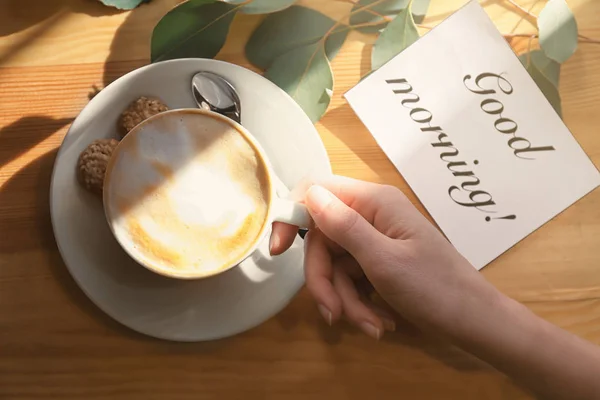 Woman holding cup of aromatic coffee at table. Tasty breakfast and card with "Good morning" wish — Stock Photo, Image