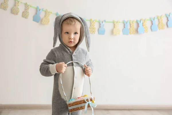 Menino bonito em traje de coelho segurando cesta com ovos de Páscoa dentro de casa — Fotografia de Stock