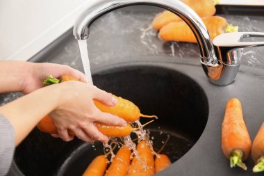 Young woman washing fresh raw carrot in kitchen clipart