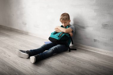 Upset little boy with backpack sitting on floor indoors. Bullying in school clipart