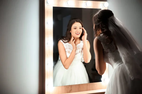 Hermosa novia joven en vestido de novia blanco con maquillaje creado por el artista profesional cerca del espejo en el interior — Foto de Stock