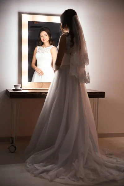 Hermosa novia joven en vestido de novia blanco con maquillaje creado por el artista profesional cerca del espejo en el interior — Foto de Stock