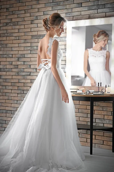 Mooie jonge bruid in witte bruiloft jurk met make-up gemaakt door professionele kunstenaar in de buurt van spiegel binnenshuis — Stockfoto