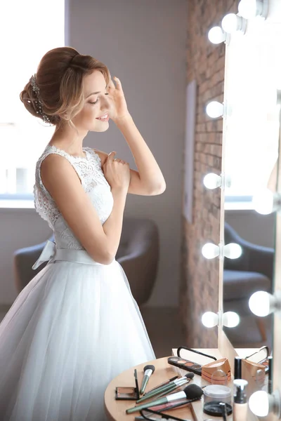 Mooie jonge bruid in witte bruiloft jurk met make-up gemaakt door professionele kunstenaar in de buurt van spiegel binnenshuis — Stockfoto