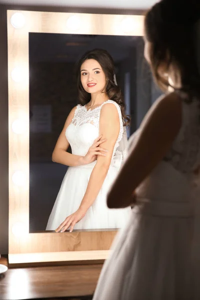 Hermosa novia joven en vestido de novia blanco con maquillaje creado por el artista profesional cerca del espejo en el interior —  Fotos de Stock