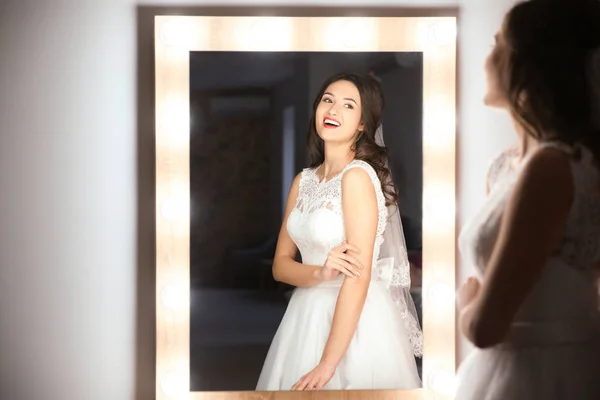 Hermosa novia joven en vestido de novia blanco con maquillaje creado por el artista profesional cerca del espejo en el interior — Foto de Stock