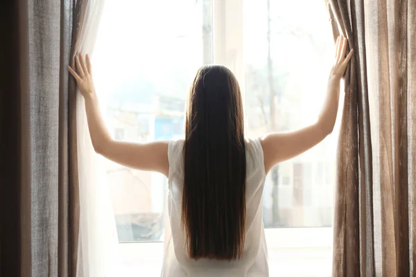 Jeune femme ouvrant rideaux à l'intérieur — Photo