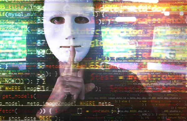 Хакер в маске показывает жест молчания и код на размытом фоне. Концепция кибератаки и безопасности — стоковое фото