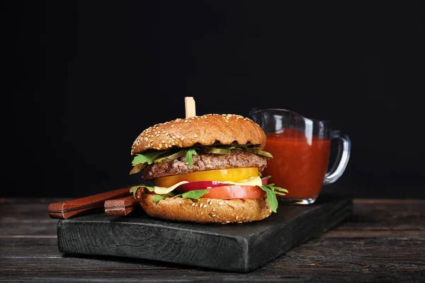 Leckere Burger mit Tomatensauce auf Holzbrett vor dunklem Hintergrund — Stockfoto