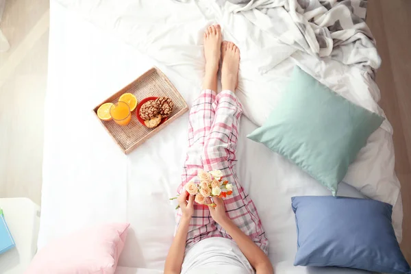 Young woman holding flowers and tray with breakfast on bed at home — Stock Photo, Image
