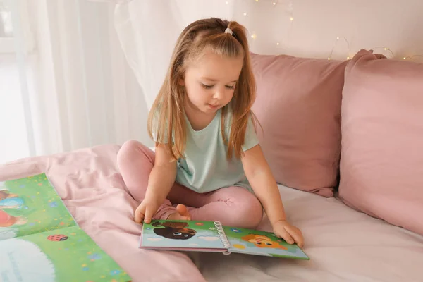 Linda niña leyendo libro mientras está sentado en la cama en casa — Foto de Stock