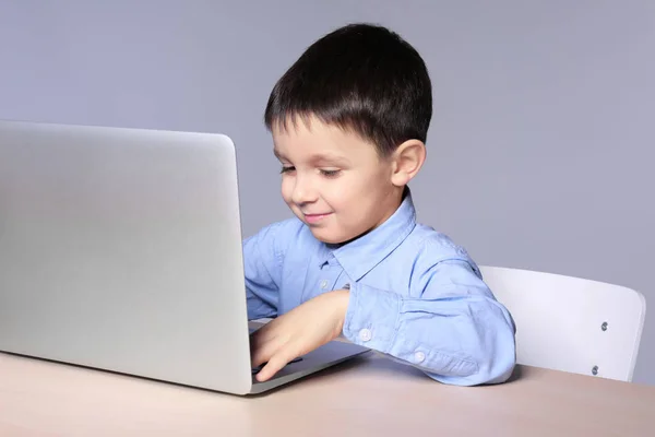 Lindo niño usando el ordenador portátil mientras hace la tarea contra el fondo gris — Foto de Stock