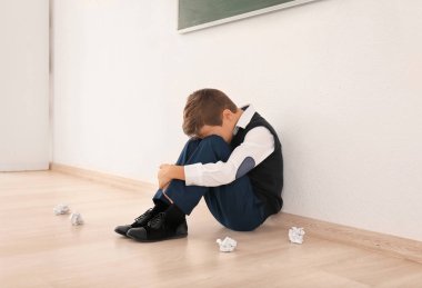 Upset little boy sitting on floor indoors. Bullying in school clipart