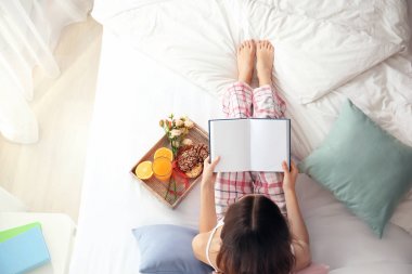 Genç kadın evde yatakta kahvaltı tepsisiyle ve kitap okuma