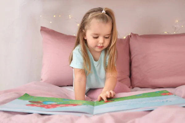 Söt liten flicka som läser bok medan du sitter på sängen hemma — Stockfoto