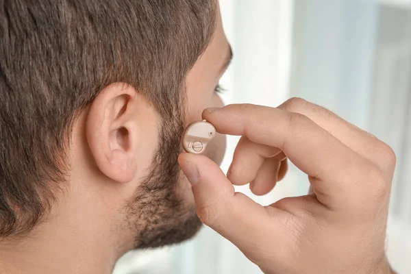 Young man putting hearing aid in ear indoors — Stock Photo, Image