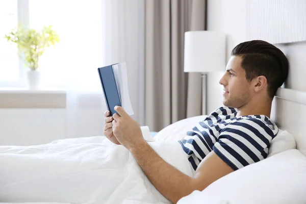 Joven hombre guapo leyendo libro en la cama en casa — Foto de Stock