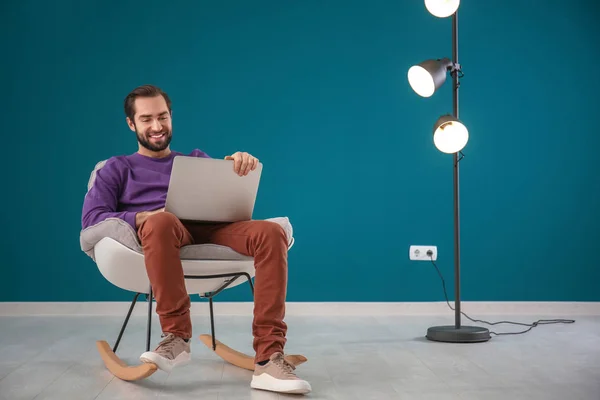 Handsome man with laptop sitting in comfortable armchair against color wall