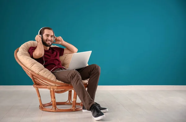Handsome man with laptop listening to music while sitting in comfortable armchair against color wall