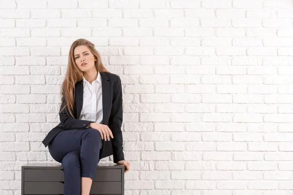 Bella donna alla moda in abito elegante seduta su cassettiera contro muro di mattoni bianchi — Foto Stock
