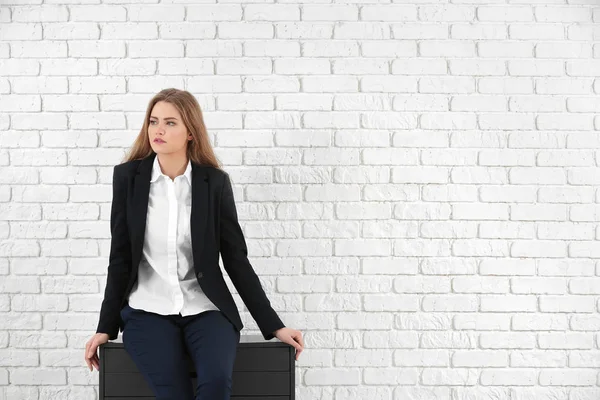 Beautiful fashionable woman in elegant suit sitting on chest of drawers against white brick wall — Stock Photo, Image