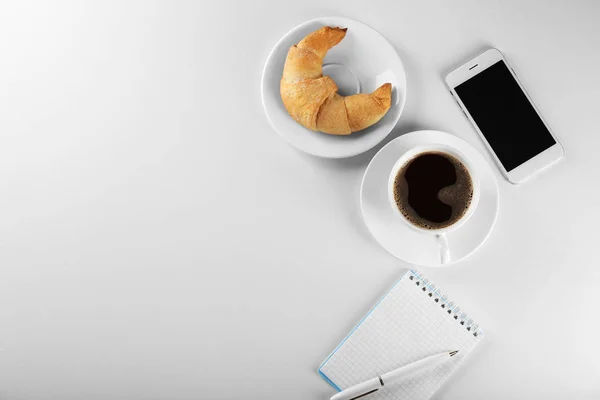 Sabroso rollo de media luna con taza de café y teléfono sobre fondo blanco —  Fotos de Stock