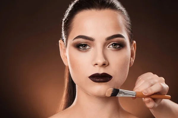 Professional visage artist applying makeup on woman's face against dark background — Stock Photo, Image