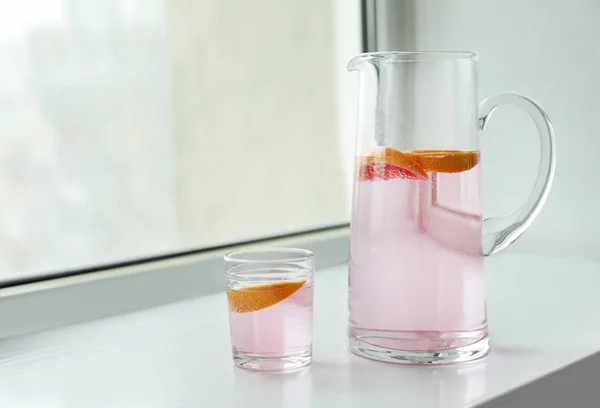 Jarra y vaso de limonada fresca con pomelo en alféizar de ventana — Foto de Stock