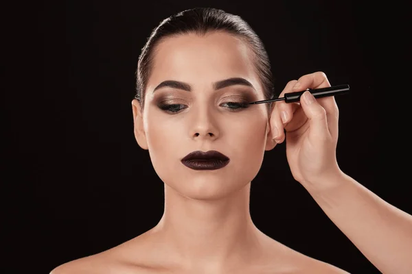 Professional visage artist applying makeup on woman's face against black background — Stock Photo, Image