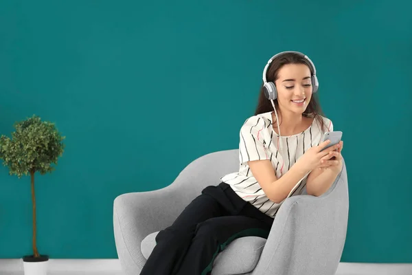 Hermosa mujer joven escuchando música mientras está sentada en un cómodo sillón contra la pared de color — Foto de Stock