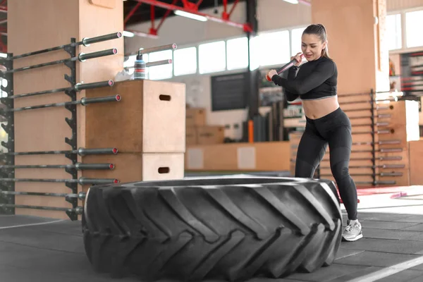 Young muscular woman hitting heavy tire with hammer in gym