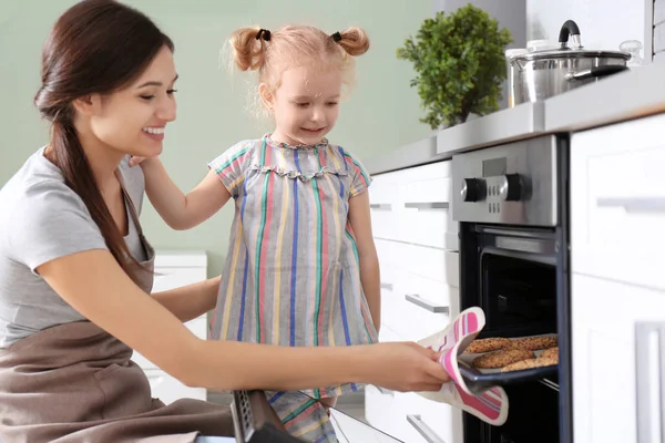 Liten flicka titta på hennes mor baka kakor i ugnen inomhus — Stockfoto