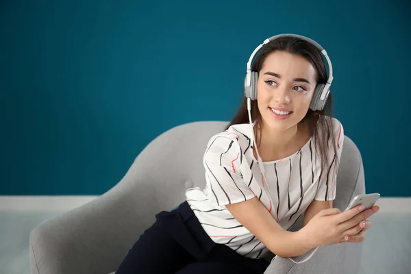 Hermosa mujer joven escuchando música mientras está sentada en un cómodo sillón contra la pared de color —  Fotos de Stock
