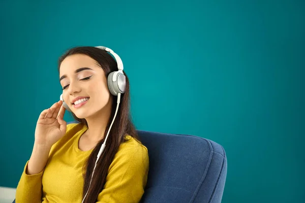 Hermosa mujer joven escuchando música mientras está sentada en un cómodo sillón contra el fondo de color —  Fotos de Stock