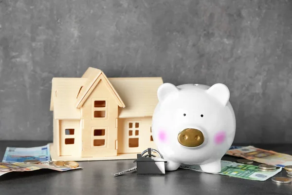 Porquinho cerâmico com dinheiro, modelo de casa e chave na mesa cinza — Fotografia de Stock
