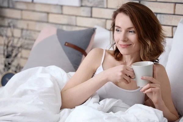 Piękna kobieta uśmiechający się filiżankę kawy, leżąc w łóżku w domu — Zdjęcie stockowe