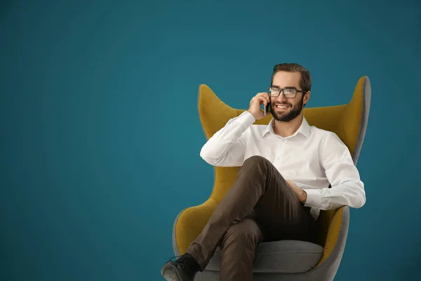 Hombre guapo hablando en el teléfono móvil mientras está sentado en un cómodo sillón contra el fondo de color —  Fotos de Stock