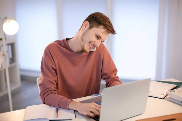 Hombre joven hablando por teléfono mientras trabaja con el ordenador portátil en el interior — Foto de Stock